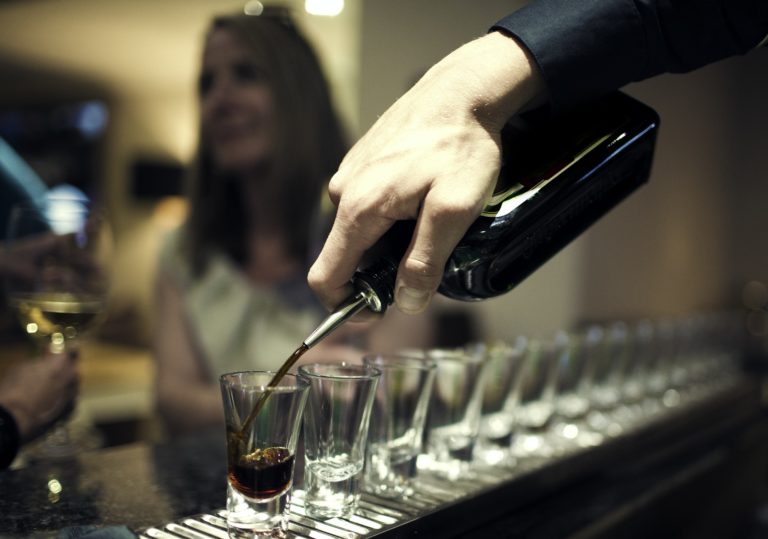 Close up of bartender pouring drinks from bottle into a row of shot glasses on bar counter.